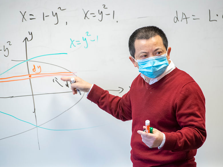 Man wearing red sweater and surgical mask points to mathematic concept on white board.