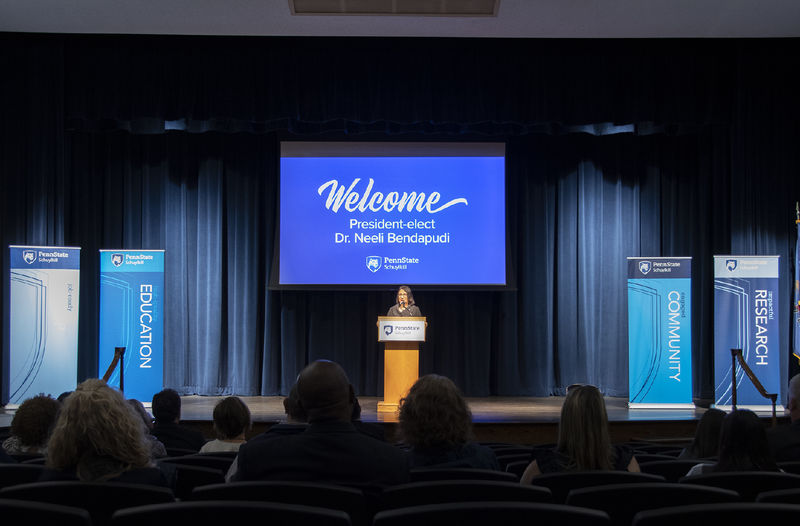 Dr. Neeli Bendapudi stands on Penn State Schuylkill's auditorium stage with blue curtains hanging from the ceiling and a welcome slide projected behind her