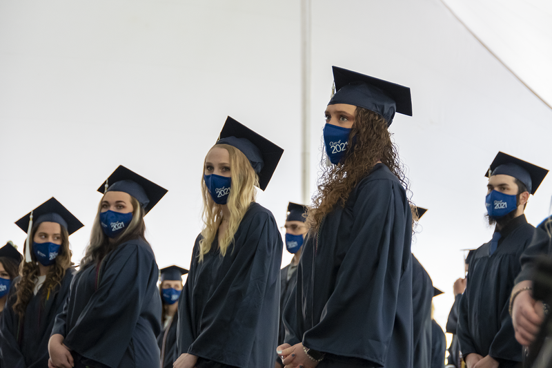 Students wearing face masks and academic regalia stand under a tent.