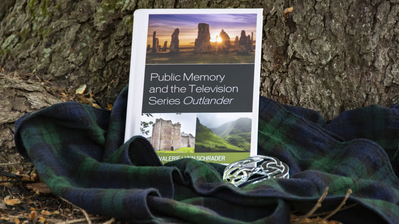 An image of the book, next to a tartan plaid scarf, leaned against a tree.