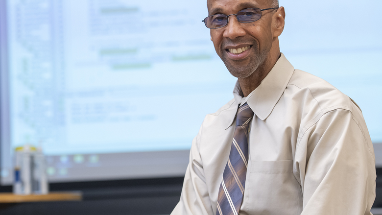 Instructor wearing button down shirt and tie sits in front of a projector with coding information on screen