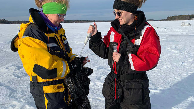 Two women wear winter weather gear and sunglasses while using a micropipette while walking on a frozen lake