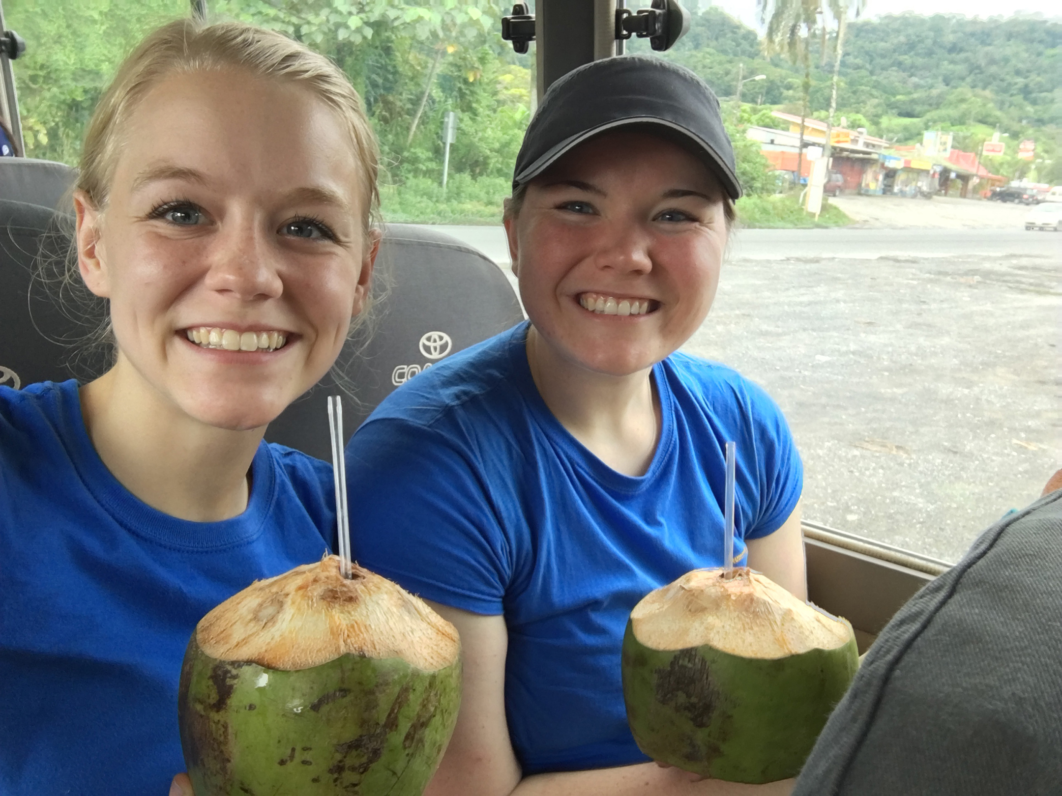 Two students sit in a chartered bus drinking from coconuts with straws coming out of the top