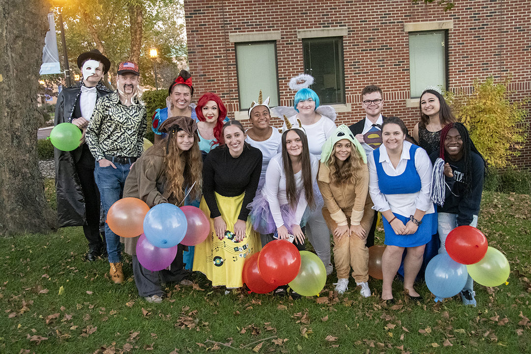 Students wearing Halloween costumes and holding balloons pose for a photo in front of a brick building