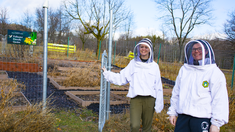 Students Marla and Alyssa stand next to the campus certified pollinator friendly garden in their bee suits.