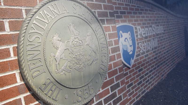 Closeup photo of campus entrance gate, a red brick wall with cast metal university seal and blue-and-white campus logo attached