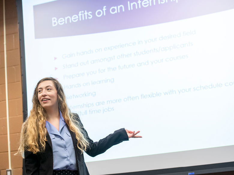 Student in business attire presenting in front of a projector screen.