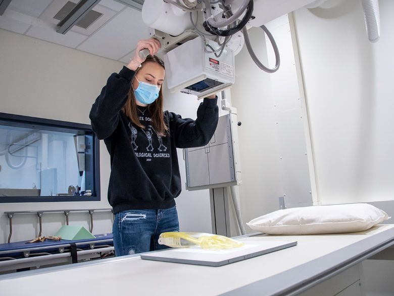 Student wearing mask and black hoodie uses X-ray machine to examine a phantom