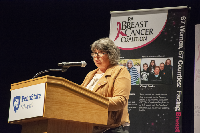Andrea Solinski, Penn State Schuylkill's lab supervisor, speaks at a podium with the Schuylkill logo adorning its front. Behind her is a panel from the PA Breast Cancer Coalition's 67 Women, 67 Counties exhibit.