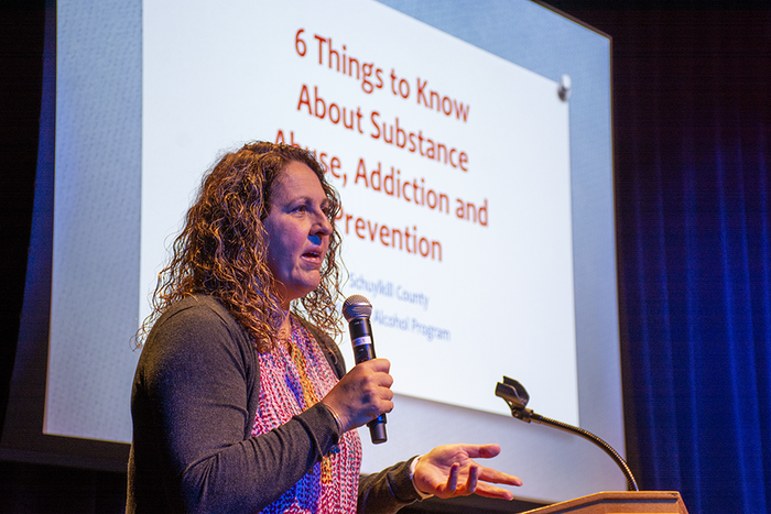 Diane Rowland holds a microphone while standing in front of a projector slide reading "Six Signs to Know about Substance Abuse, Addiction, and Prevention
