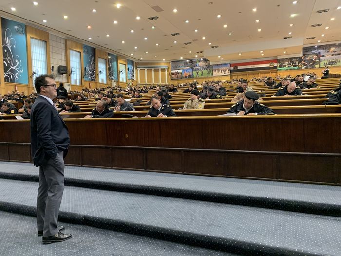 Dr. Hakan Can stands at the foot of a room with auditorium-style seating while uniformed police officers fill out a survey on paper