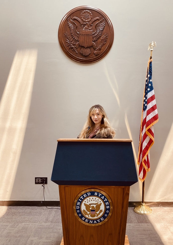 An image of student-intern Corrine Ellis standing at a podium with a U.S. Senate seal.