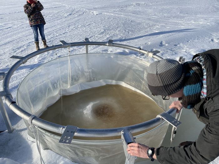 People wearing winter gear stand around a hole in a frozen lake. A metal circle with hanging plastic helps the researchers isolate samples from the lake.