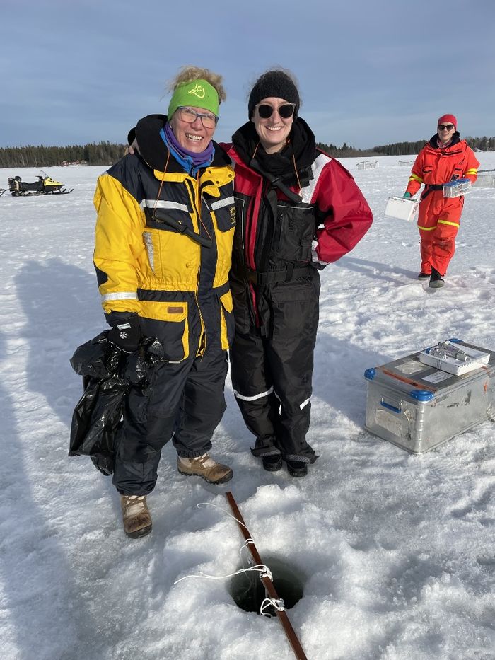 Two women wearing winter weather gear pose for a photo on a frozen lake. There is a hole in the ice in front of them with scientific equipment surrounding it.