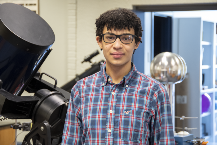 Student wearing glasses and a plaid shirt standing in front of a telescope and other physics-related lab equipment.