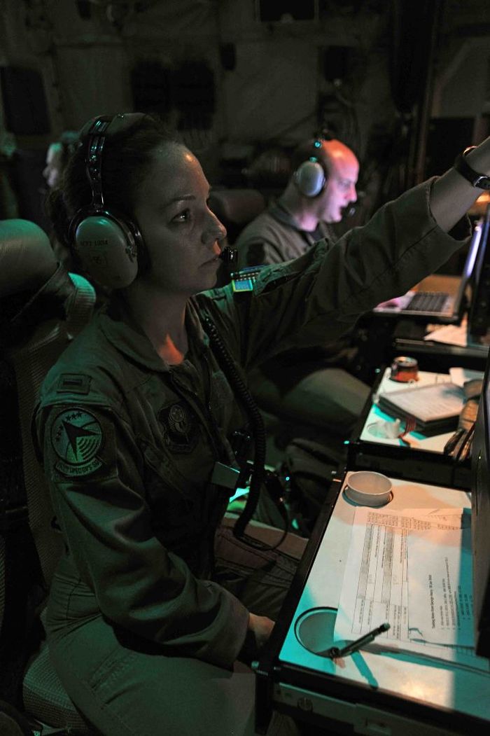 A woman in the armed forces wears headphones and points at a computer screen in a dimly lit room.