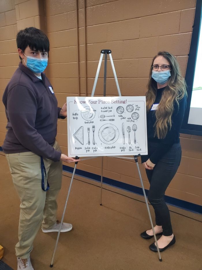 Two students wearing masks present a poster of a proper dinner setting