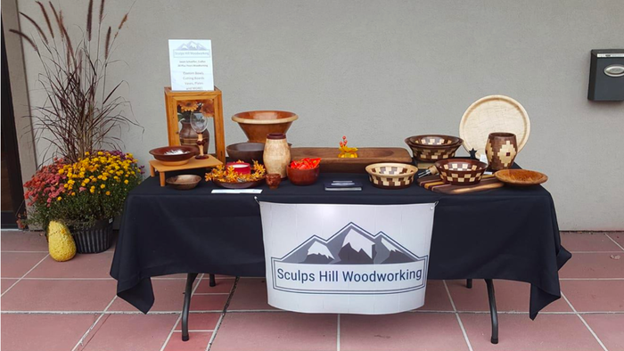 Photo of craft fair vendor table with black table cloth, sign that reads "Sculps Hill Woodworking," and several examples of the bowls and charcuterie boards they offer.