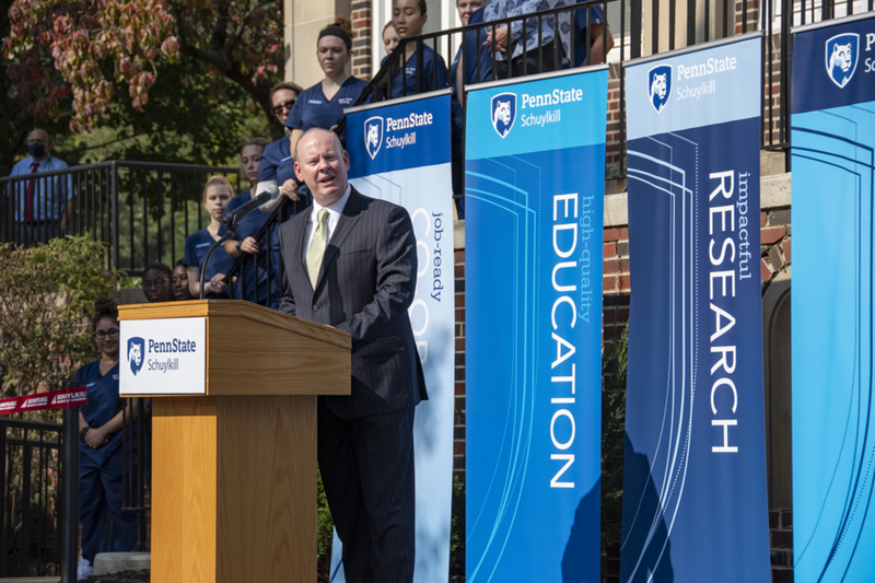 Chancellor Patrick M. Jones wears a pinstripe suit and addresses a crowd from a wooden podium with Penn State Schuylkill logo