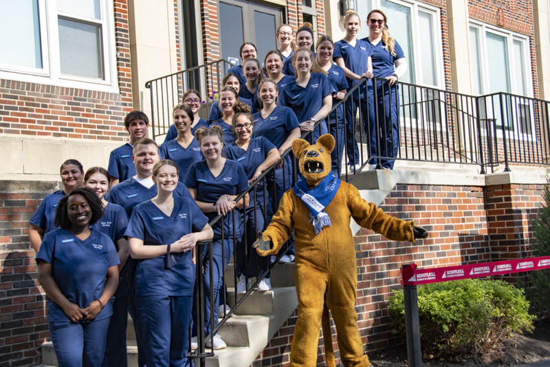 Student nurses wearing navy blue scrubs stand on stairs outside of a brick building with the Nittany Lion mascot