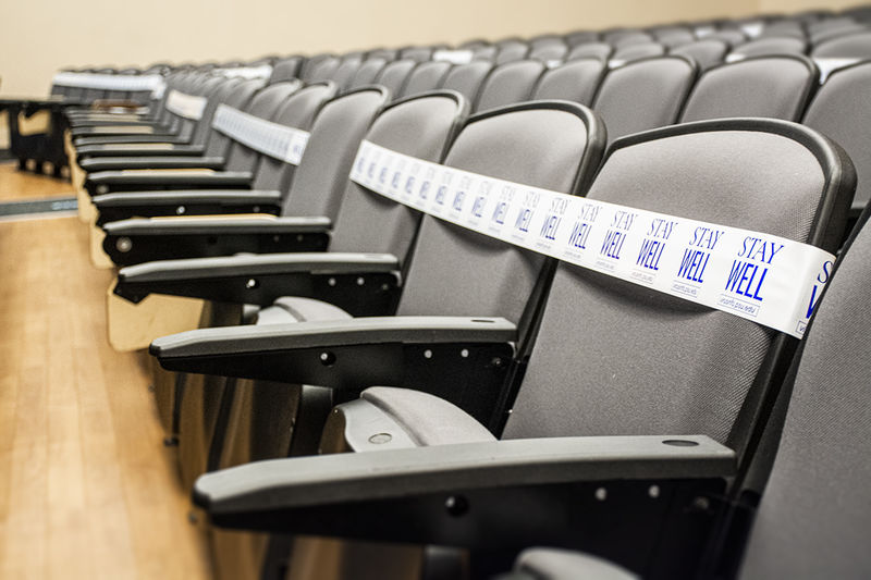 A line of gray auditorium seats. Every three chairs are blocked off with white tape reading "STAY WELL / virusinfo.psu.edu"