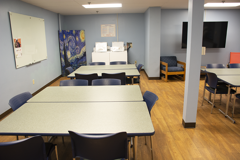 Three tables with six chairs each sit on hard wood floors. A washer and dryer are on the far end of the room. A large replica of Van Gogh's 'Starry Night' stands next to a large white board mounted to the wall. A large TV monitor is mounted to an adjacent wall.