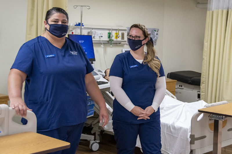 Two nursing students wearing navy scrubs and face masks show off the clinical skills nursing lab.