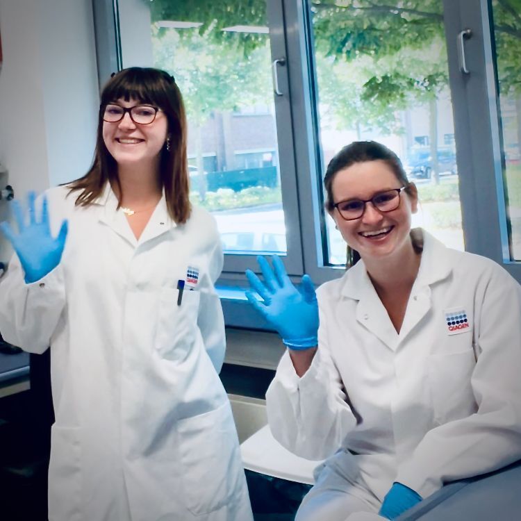 Two students wearing lab coats and blue rubber gloves smile for the camera