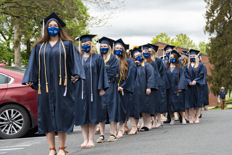 Students wearing academic regalia and face masks stand in a line in a parking lot