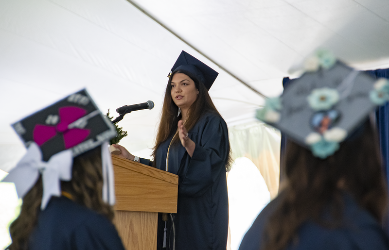 A student wearing academic regalia stands and speaks into a microphone at a podium