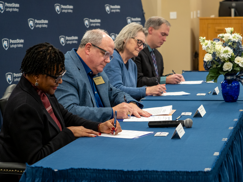 four individuals sit at a long table with their heads down and sign a document 