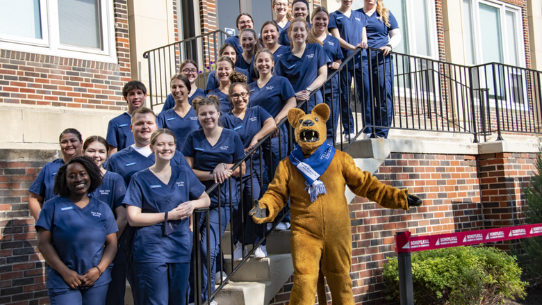 Student nurses wearing navy blue scrubs stand on stairs outside of a brick building with the Nittany Lion mascot