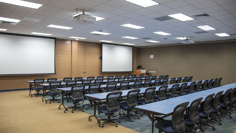 Room with brown walls and two projector screens hanging from drop ceiling with long blue tables and black chairs on wheels