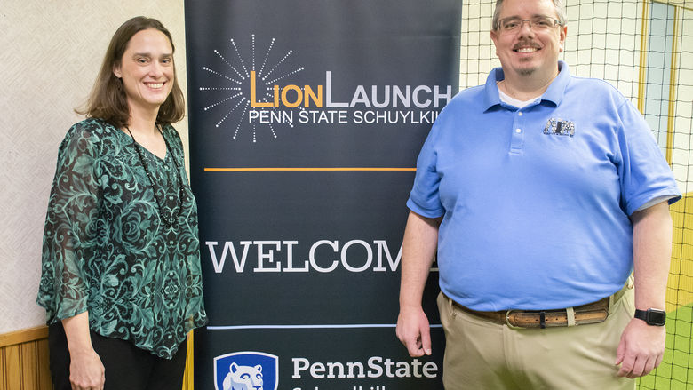 Darcy Medica, left, stands with her husband Joe, right, with a "WELCOME" sign between them featuring Penn State Schuylkill and Penn State Schuylkill LionLaunch logos.