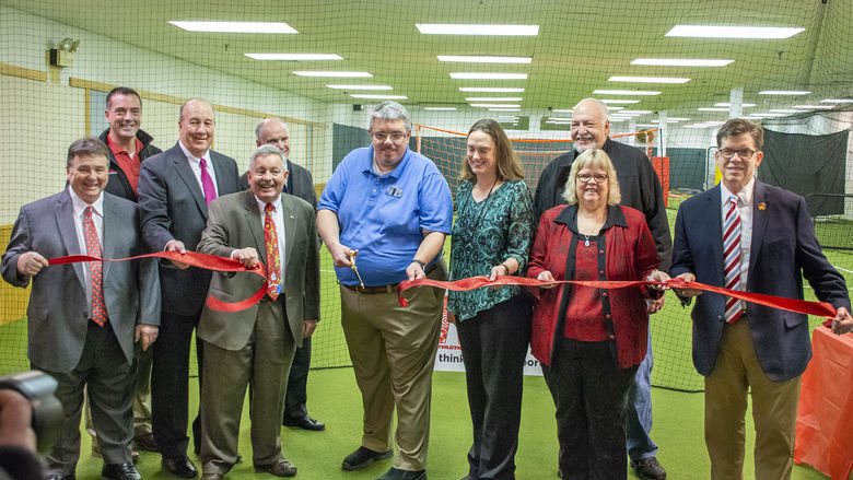 Several people stand on turf grass in a line while holding a red ribbon. Joe Medica stands in the center holding scissors to cut the ribbon.