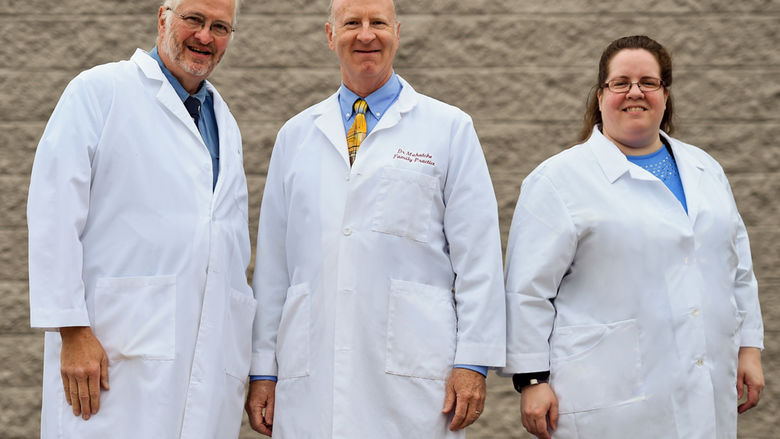 Three people -- two men and one woman -- stand in front of a beige, textured wall, posing for a photo. They're each wearing white clinical jackets.