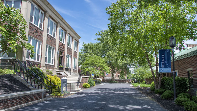 Two story brick building on left hand of photo with concrete pathway lined by trees