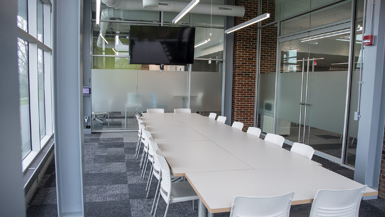 Room with floor-to-ceiling glass windows, steel support beams, and long white tables with white chairs overlooking a large monitor screen