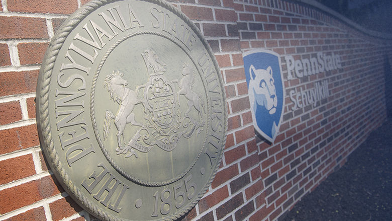 Closeup photo of campus entrance gate, a red brick wall with cast metal university seal and blue-and-white campus logo attached