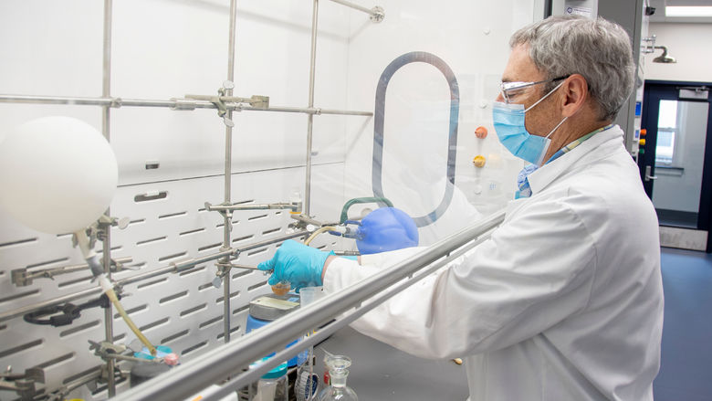 Man wearing lab coat, latex gloves, mask, and protective goggles pours a liquid into another canister in an organic chemistry lab