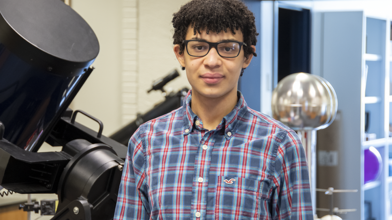 Student wearing glasses and a plaid shirt standing in front of a telescope and other physics-related lab equipment.