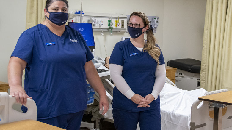 Two nursing students wearing navy scrubs and face masks show off the clinical skills nursing lab.
