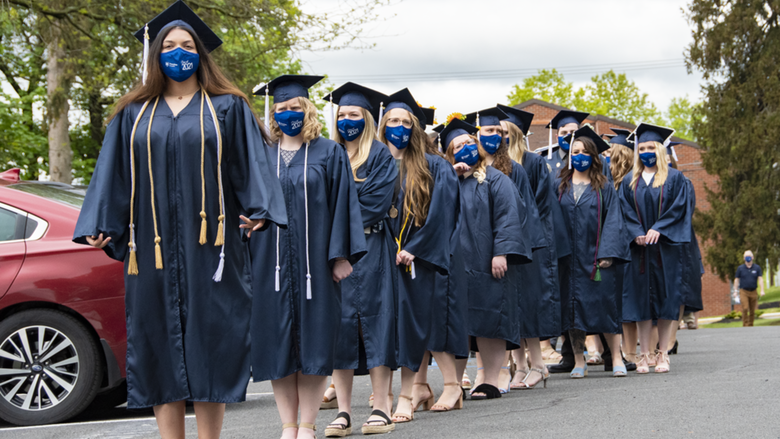 Students wearing academic regalia and face masks stand in a line in a parking lot