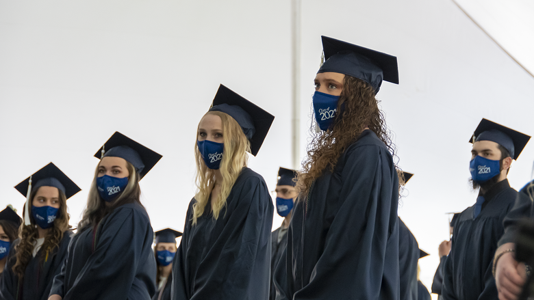 Students wearing face masks and academic regalia stand under a tent.