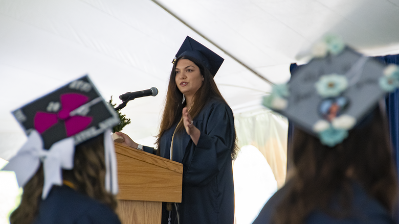 A student wearing academic regalia stands and speaks into a microphone at a podium