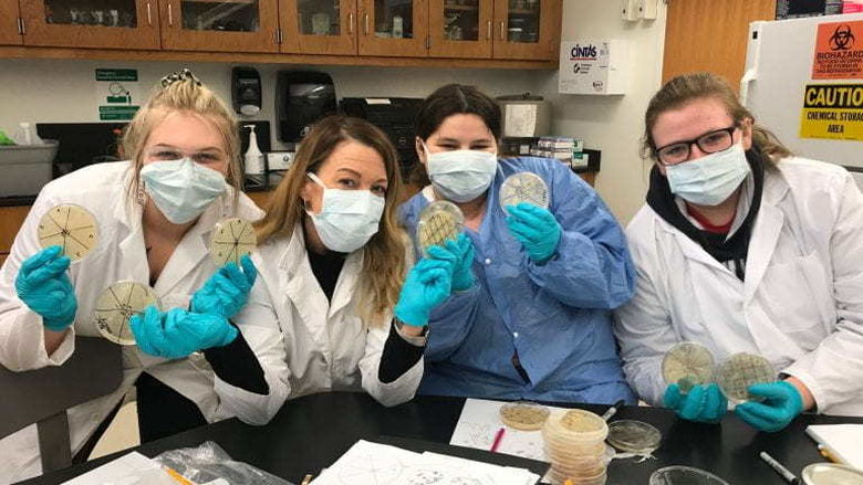 Four students wearing gloves, face masks, and lab coats hold up petri dishes smeared with diluted soil samples
