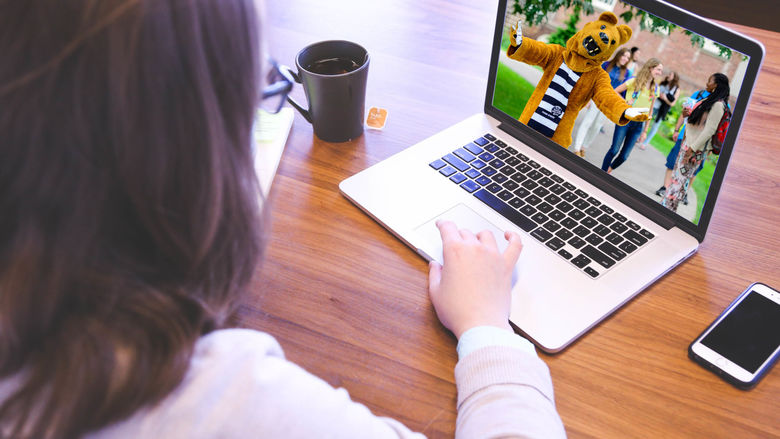 Girl sitting behind laptop with a mug of hot tea and a cell phone sitting next to her. Penn State's Nittany Lion mascot welcomes her on the laptop screen.