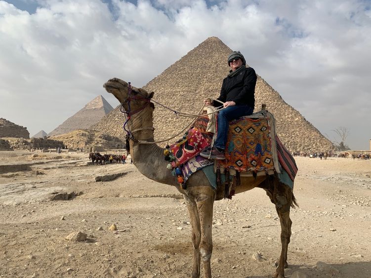 Dr. Hakan Can sits atop a camel with three pyramids behind him