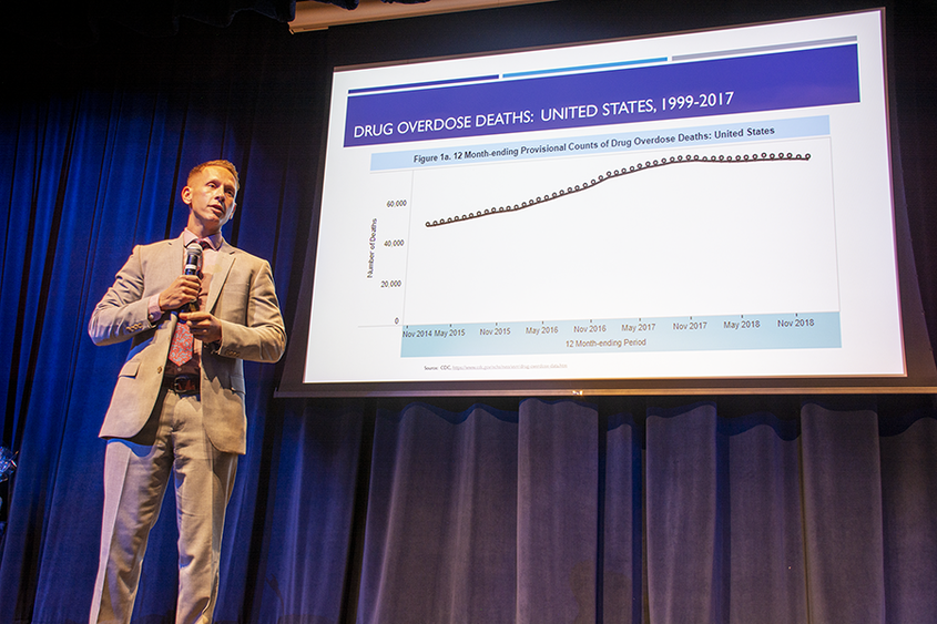 Dr. Glenn Sterner stands on stage as he delivers a speech. There is a slide behind him reviewing drug overdose deaths in the U.S. from 1999-2017 