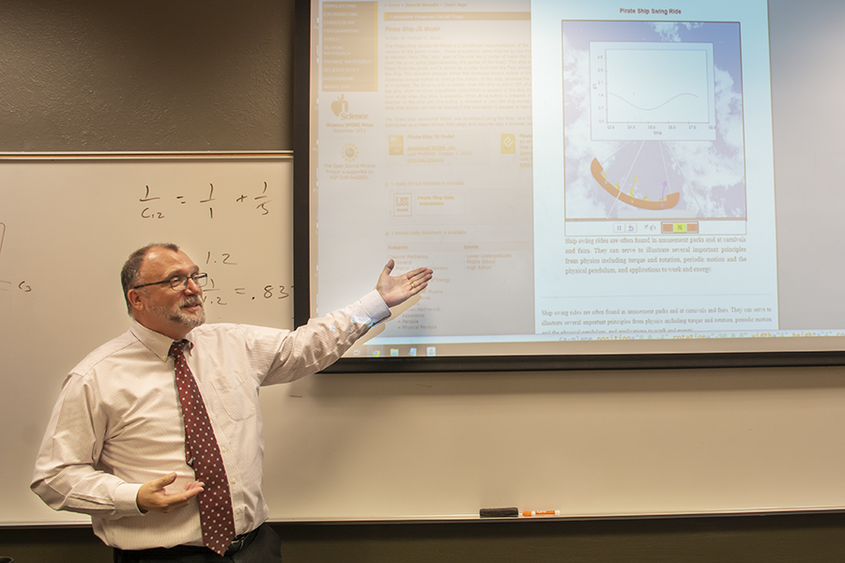 Michael Gallis stands in front of a white board with fractions written on it as he points to a projection screen showing the physics of an amusement park's pirate ship ride.